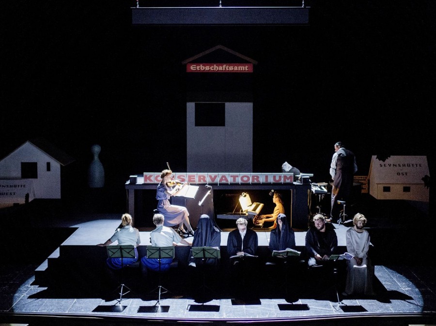 Image of konservatorium with a spotlight on a violin and piano player with 7 individuals at the foot of the stage