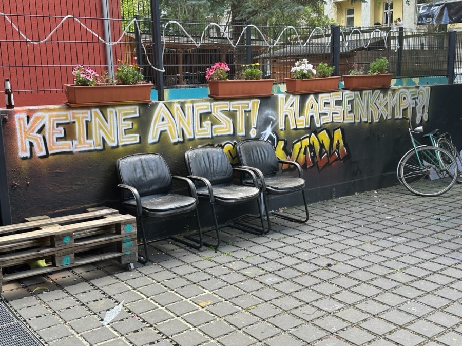 Wall with grafitti in German language with 3 chairs infront, and several flower boxes on the wall.